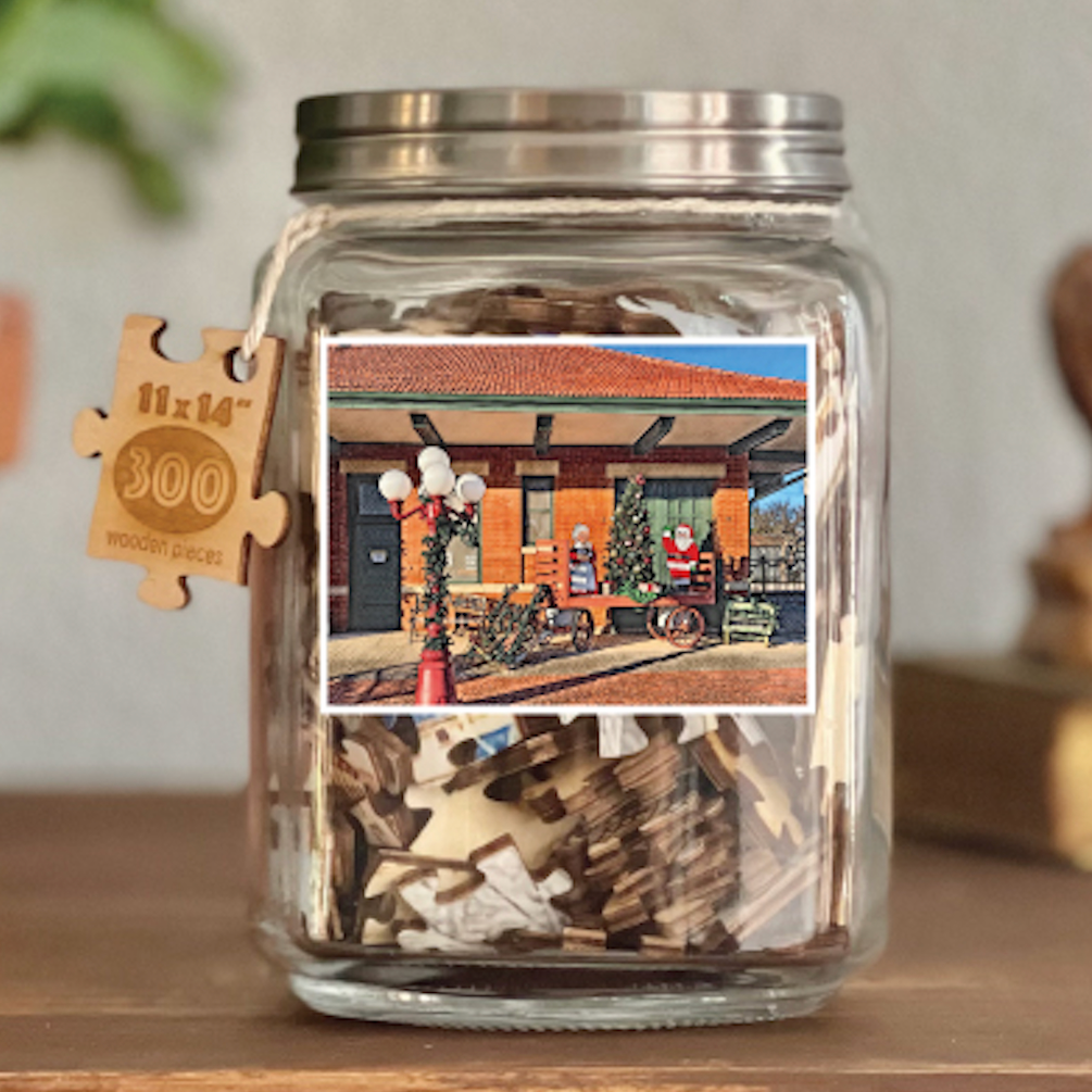 Image of puzzle shown on display clear glass Mason style jar with silver metal crew on lid. Exposes puzzle pieces inside the jar. Wooden tag hangs down the side of the jar with kite string from lid of jar with size and piece count engraved into it.