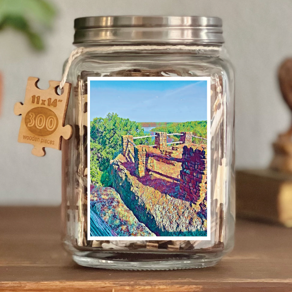 Image of puzzle shown on display clear glass Mason style jar with silver metal crew on lid. Exposes puzzle pieces inside the jar. Wooden tag hangs down the side of the jar with kite string from lid of jar with size and piece count engraved into it.