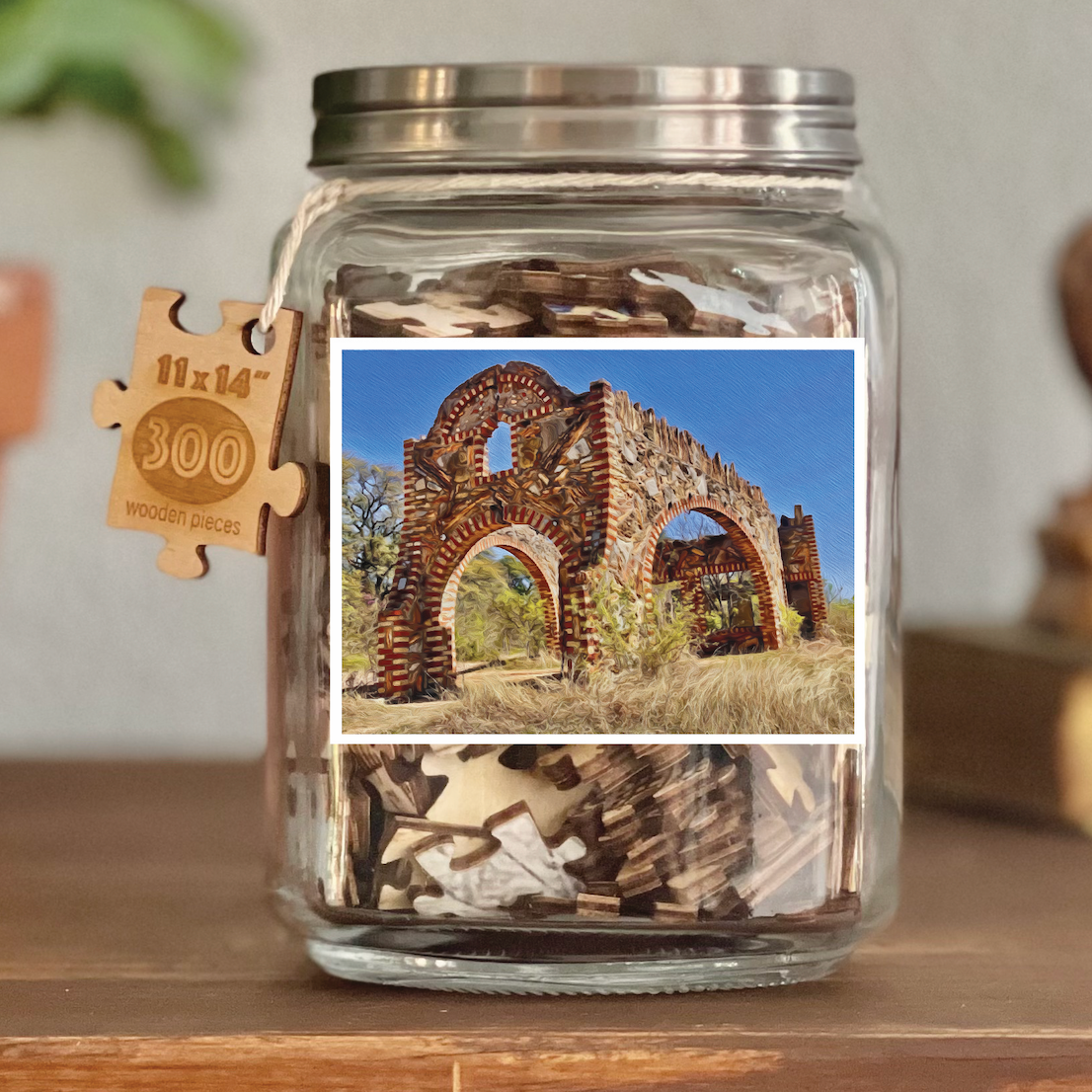 Image of puzzle shown on display clear glass Mason style jar with silver metal crew on lid. Exposes puzzle pieces inside the jar. Wooden tag hangs down the side of the jar with kite string from lid of jar with size and piece count engraved into it.