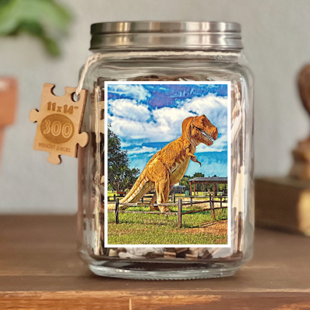 Image of puzzle shown on display clear glass Mason style jar with silver metal crew on lid. Exposes puzzle pieces inside the jar. Wooden tag hangs down the side of the jar with kite string from lid of jar with size and piece count engraved into it.