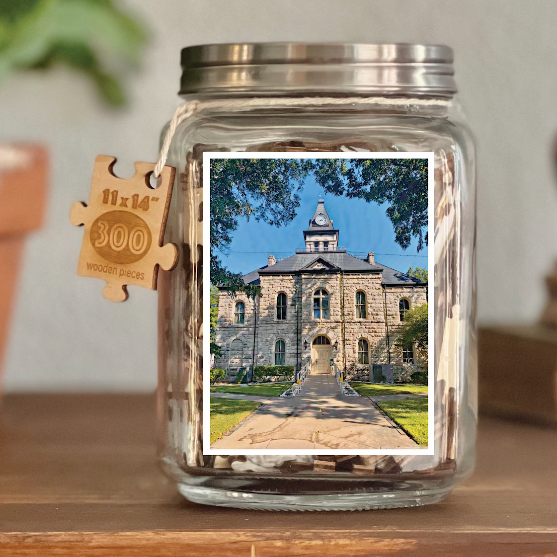 Image of puzzle shown on display clear glass Mason style jar with silver metal crew on lid. Exposes puzzle pieces inside the jar. Wooden tag hangs down the side of the jar with kite string from lid of jar with size and piece count engraved into it.